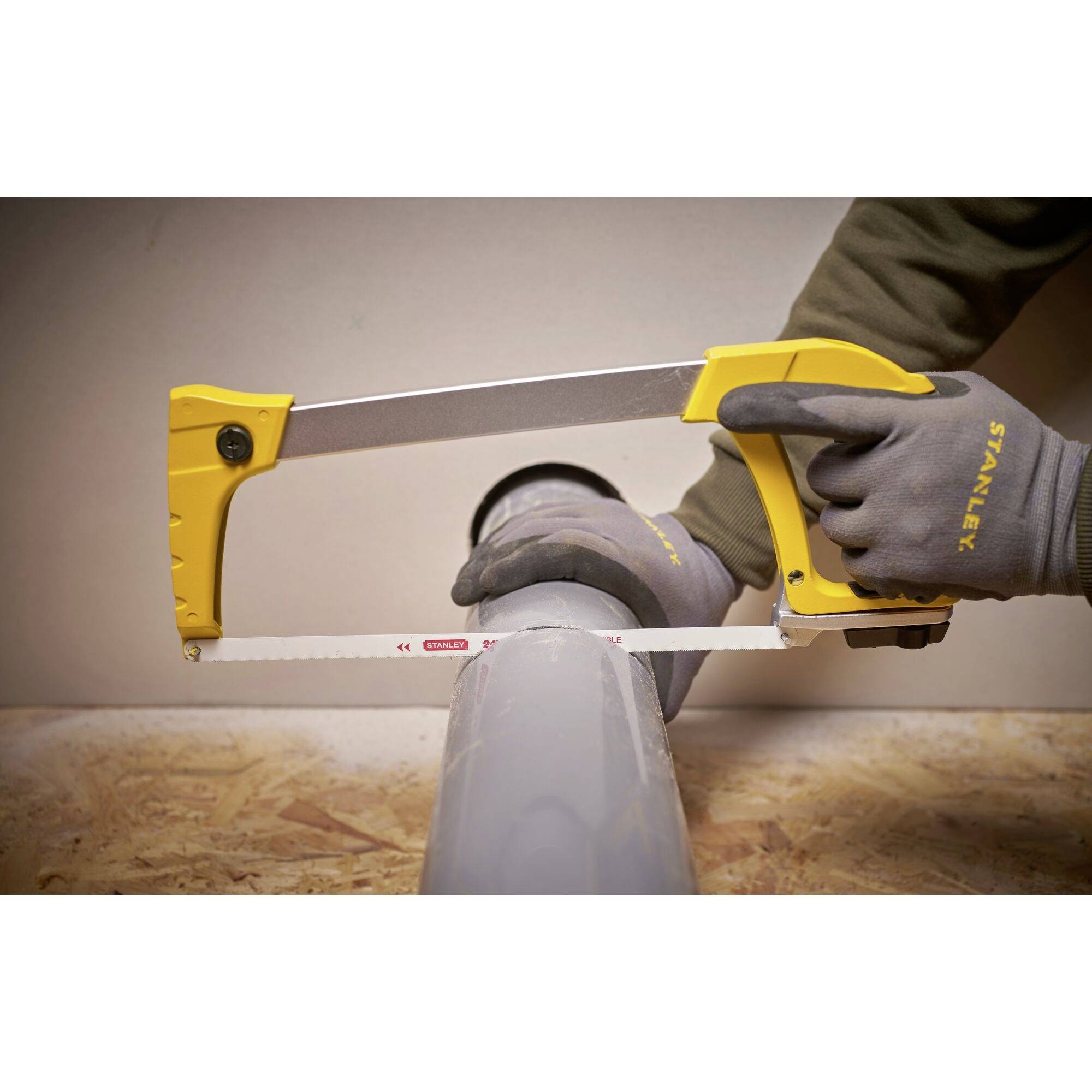 A person is sawing a grey pipe with a yellow handsaw. They are wearing grey gloves. The surface is made of wood.