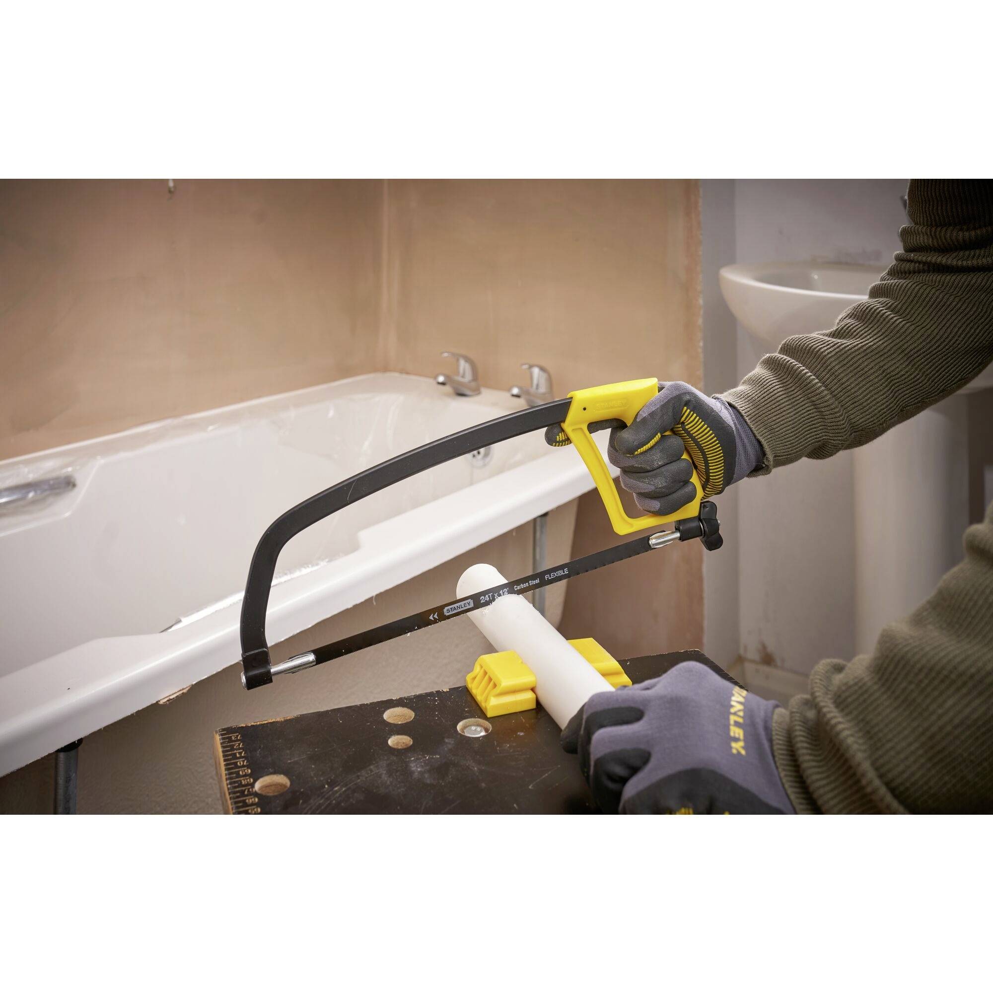 A person is cutting a white pipe with a metal saw secured in a yellow mount. A bathtub is visible in the background.