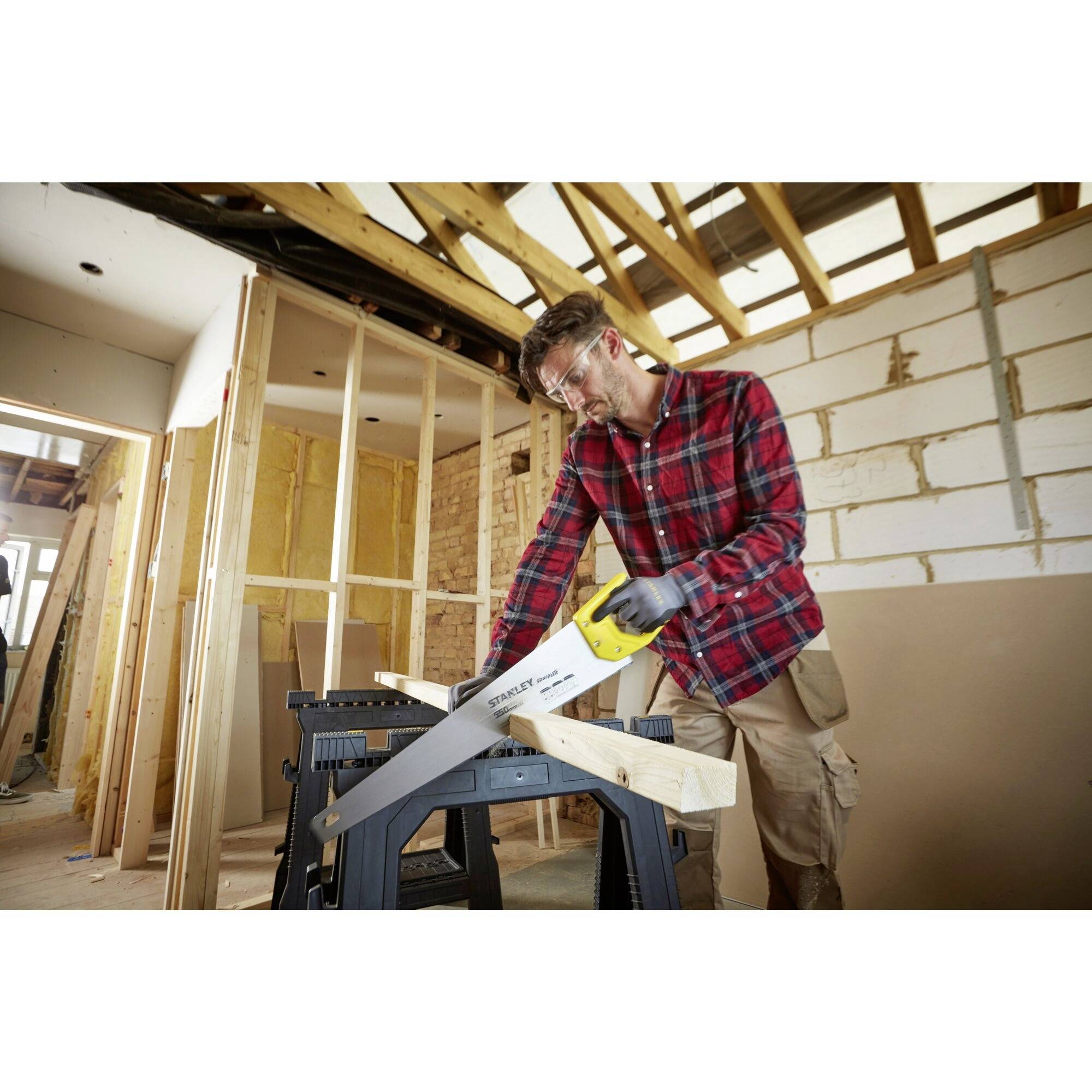 A person in work attire is using a reciprocating saw to cut wood in an unfinished room. Incomplete walls are visible in the background.