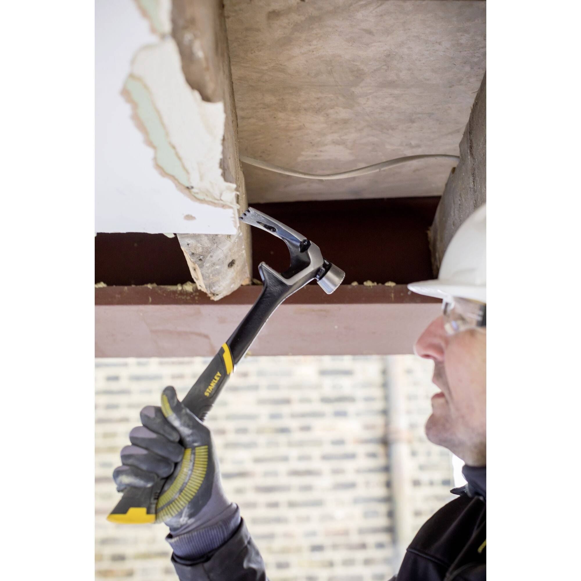 A construction worker wearing a hard hat is using a crowbar to remove a plank from a beam. A brick wall is visible in the background.
