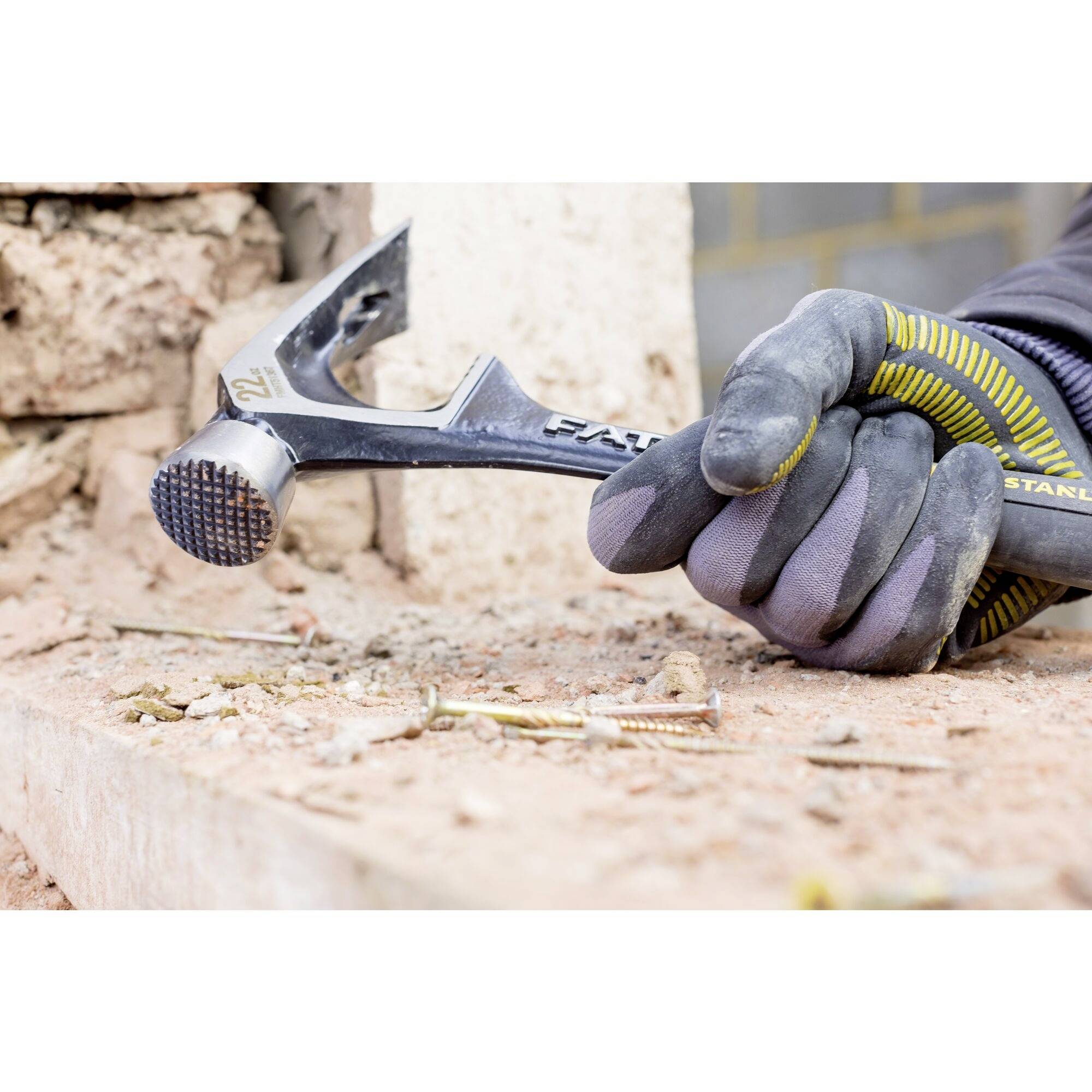 A person wearing gloves is hammering a nail into a brick wall. The focus is on the hammer and the wall.