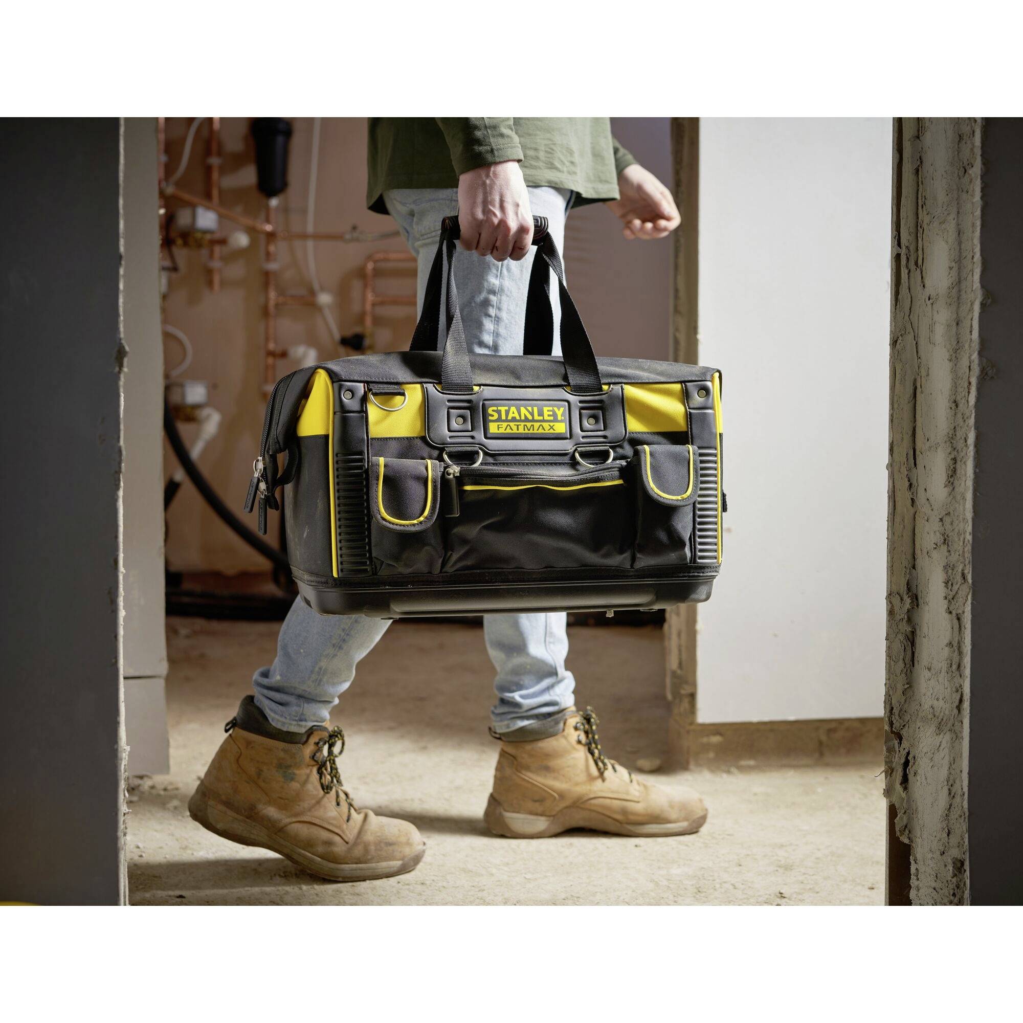 A person is carrying a tool bag. The bag is yellow and black with a 'Stanley' logo. Pipes can be seen in the background.