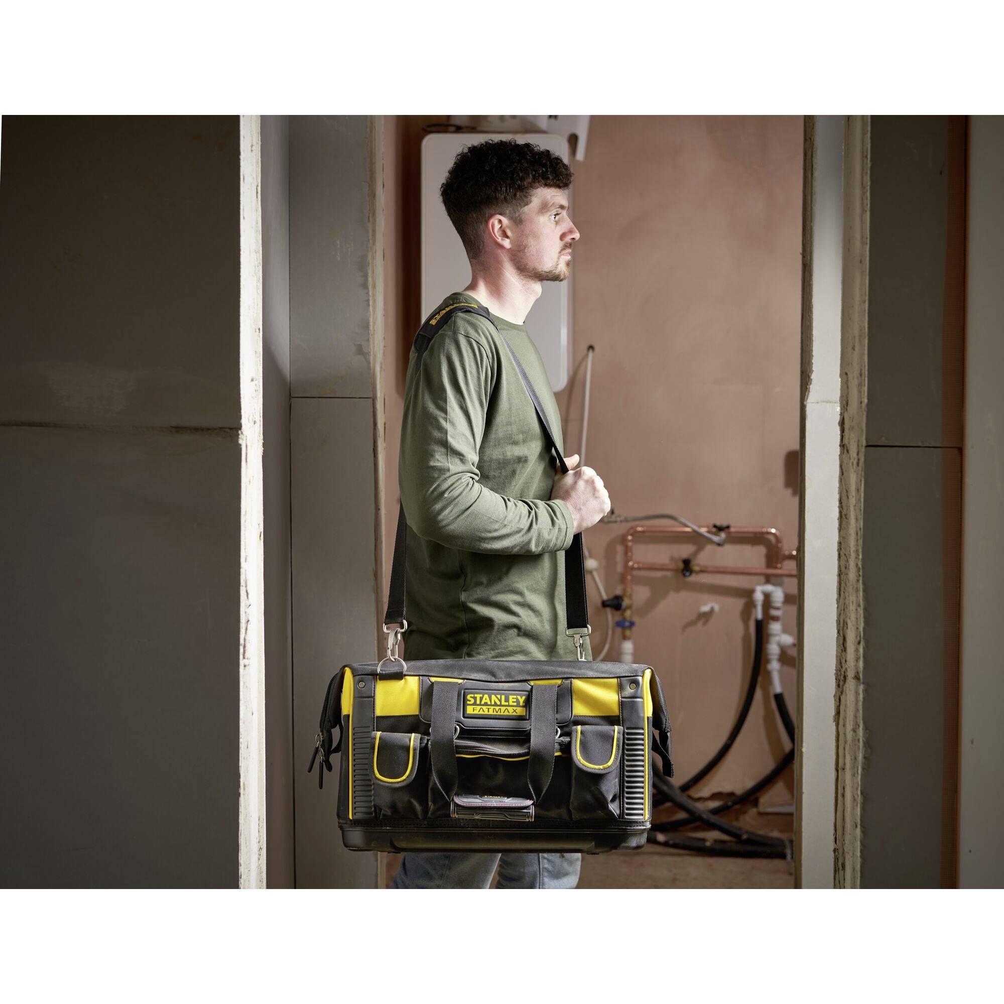 A man stands in an unfinished room, carrying a large yellow and black Stanley tool bag, prepared for craftwork.