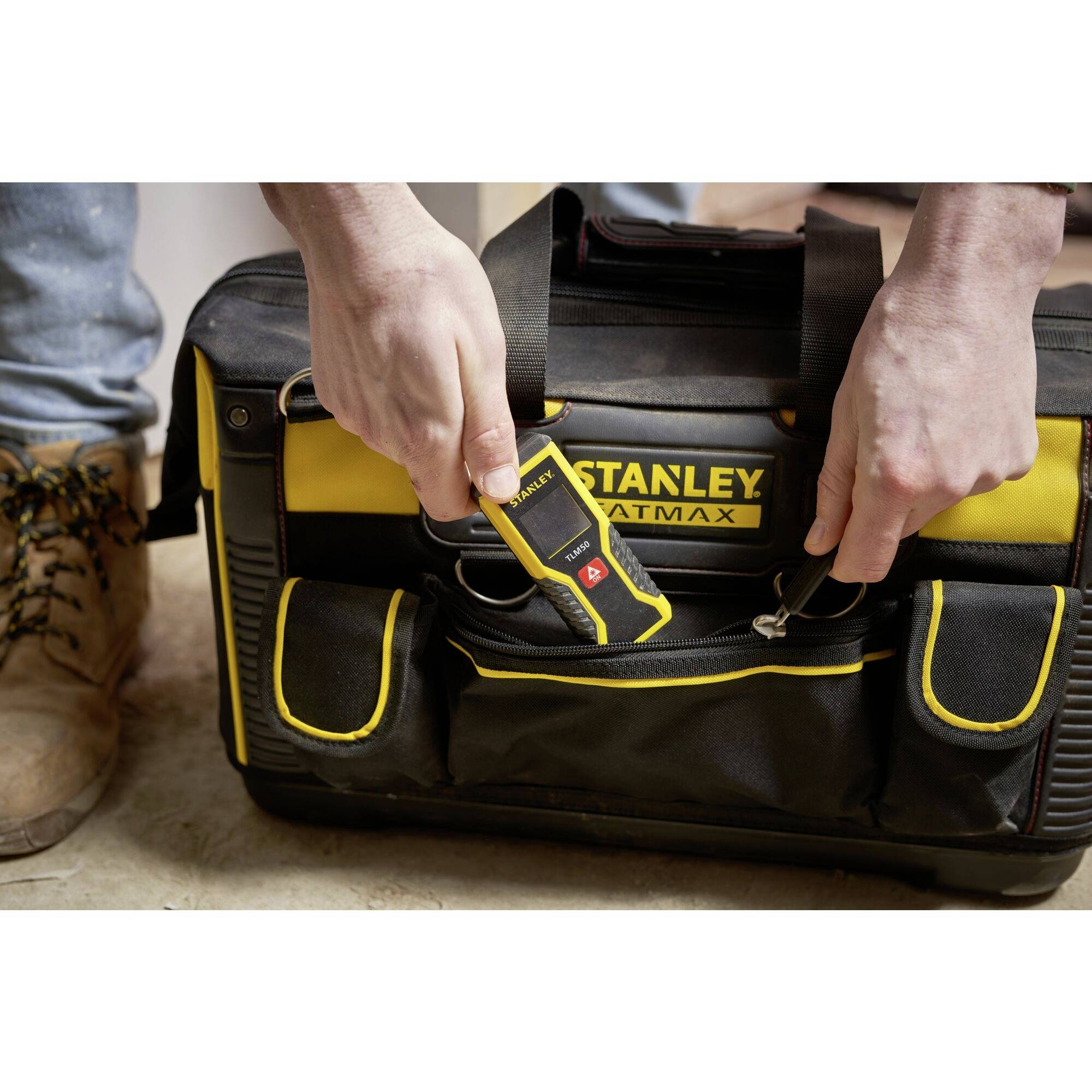 A person reaches for a yellow measuring device from a black tool bag. The bag bears the 'Stanley' brand.
