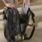 A person is placing a laptop into a black tool backpack on a construction site. A tape measure lies in the foreground.