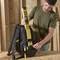 A man places a spirit level on the back of a black rucksack. Wooden walls can be seen in the background.