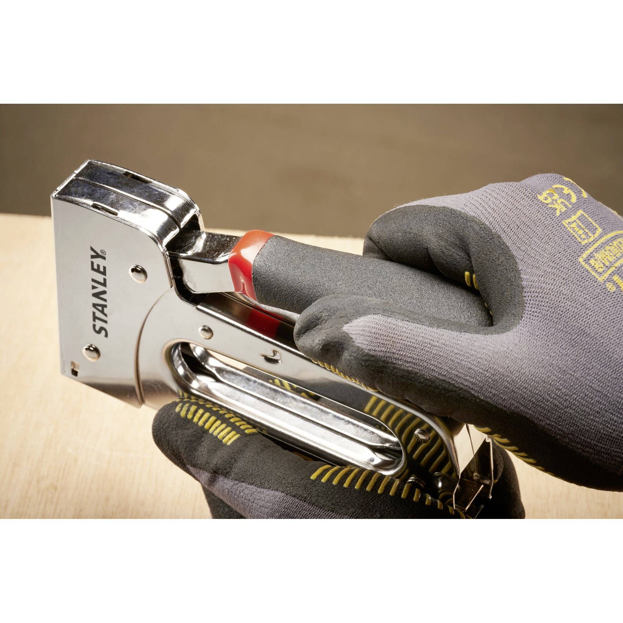 A hand wearing a grey glove holds a silver staple gun over a wooden board, ready to staple.