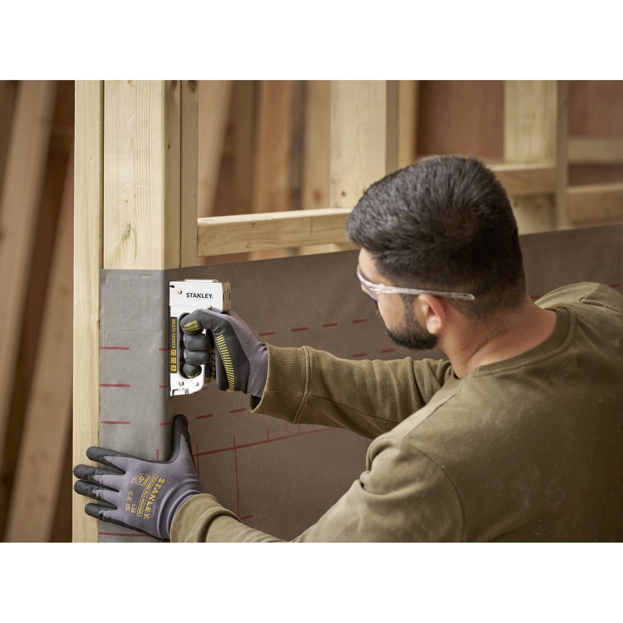A man is working on a wooden frame and using a tool to measure or mark. He is wearing safety glasses and gloves.