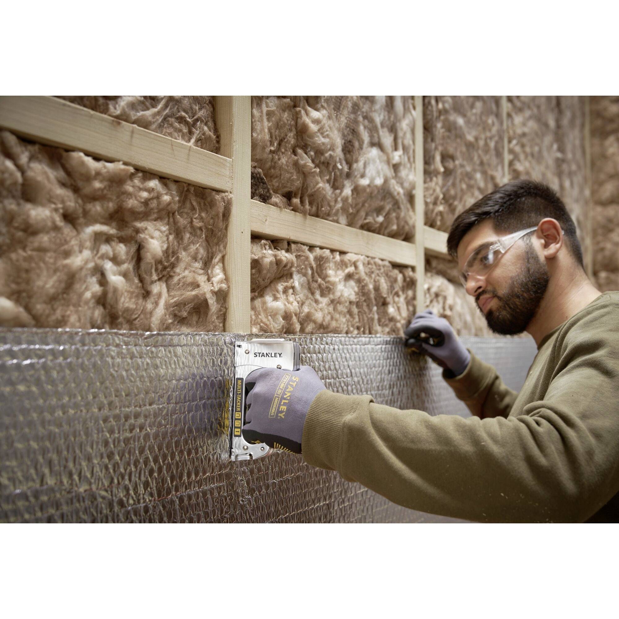 A man wearing safety glasses is measuring an insulated wall with a tape measure. The wall has an insulation layer of fibreglass.