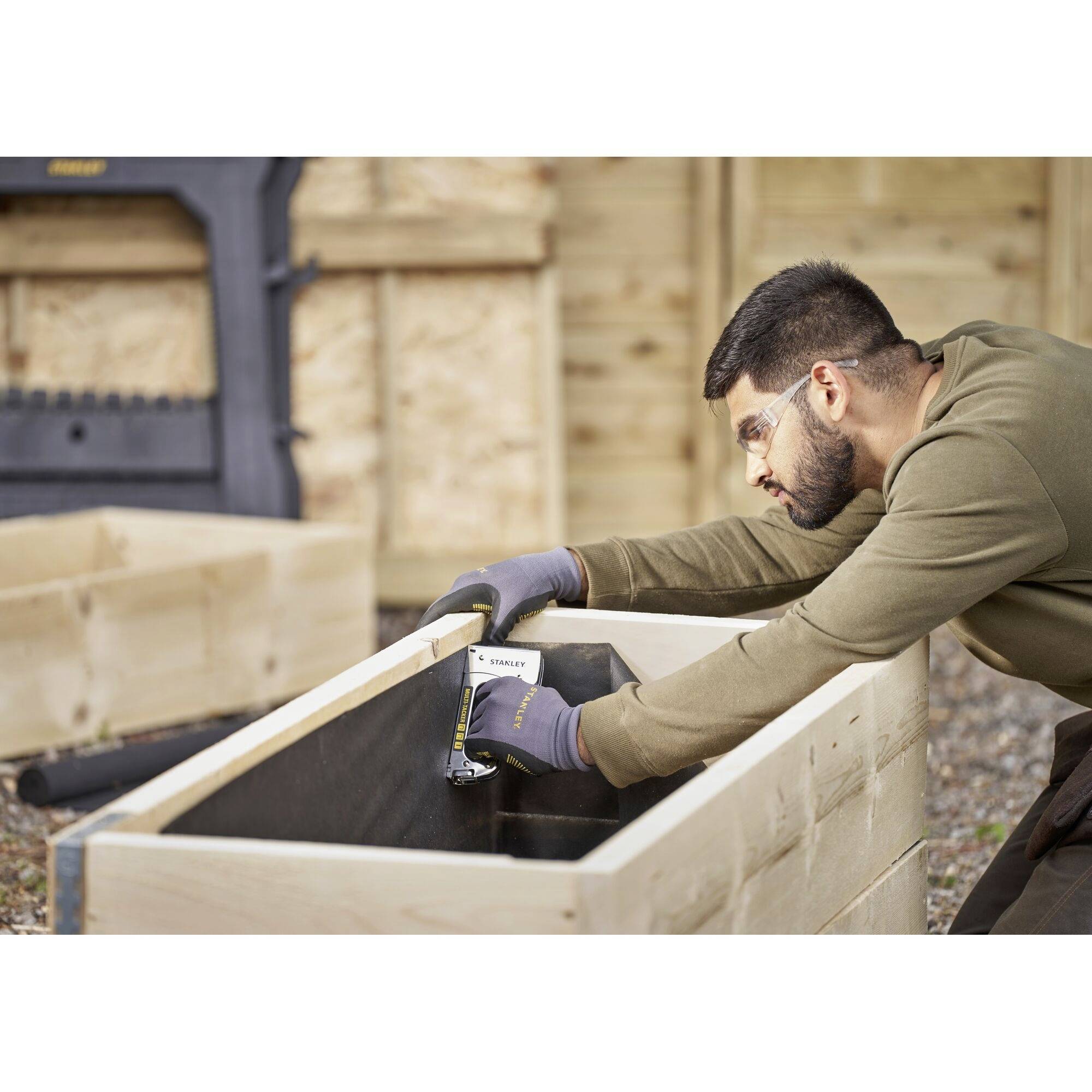 A man is using a tape measure to measure a wooden box in a workshop. He is wearing safety glasses and gloves.