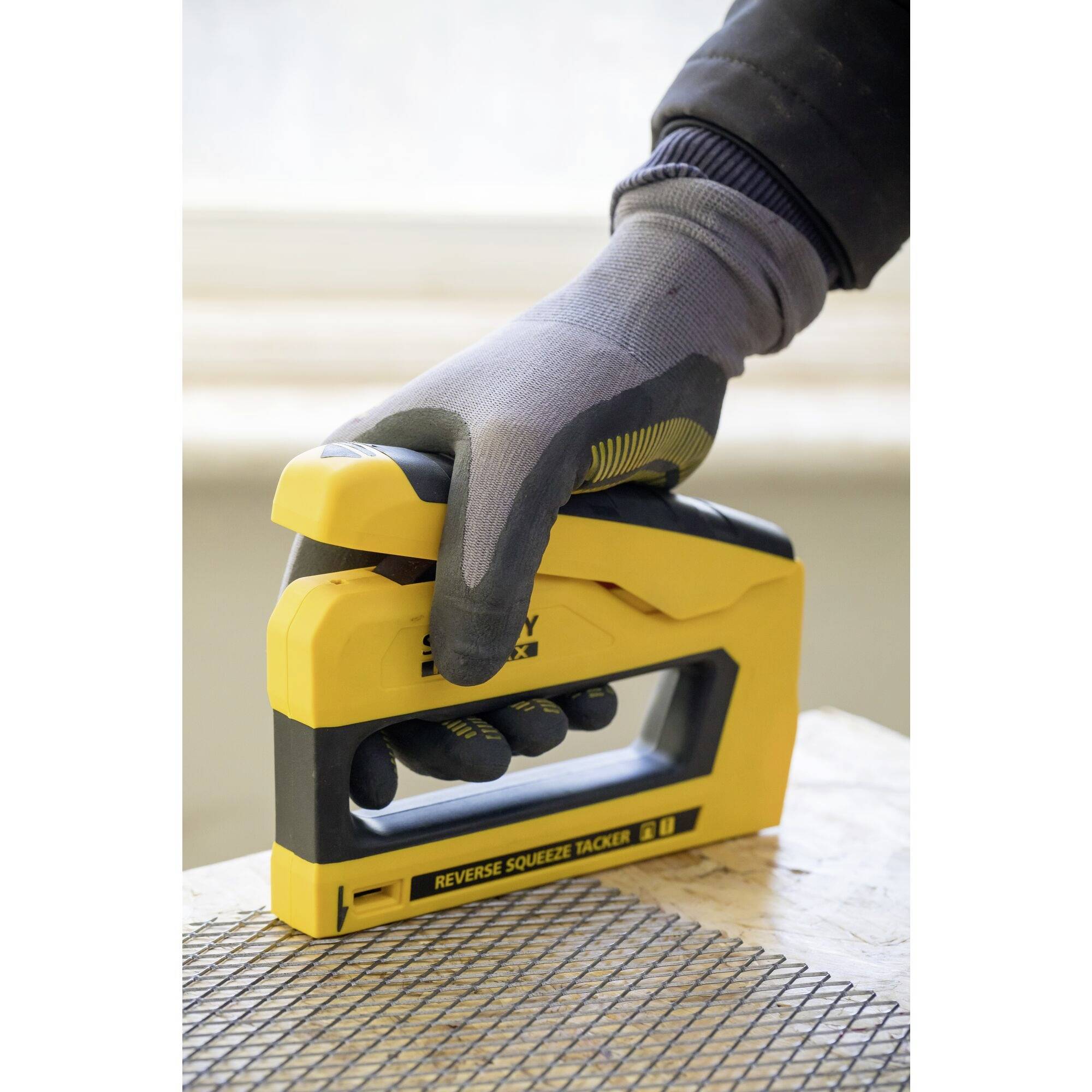 A hand in a glove holds a yellow staple gun over a wire grid on a table. The staple gun bears the text 'Reverse Squeeze Tacker'. A window is visible in the background.