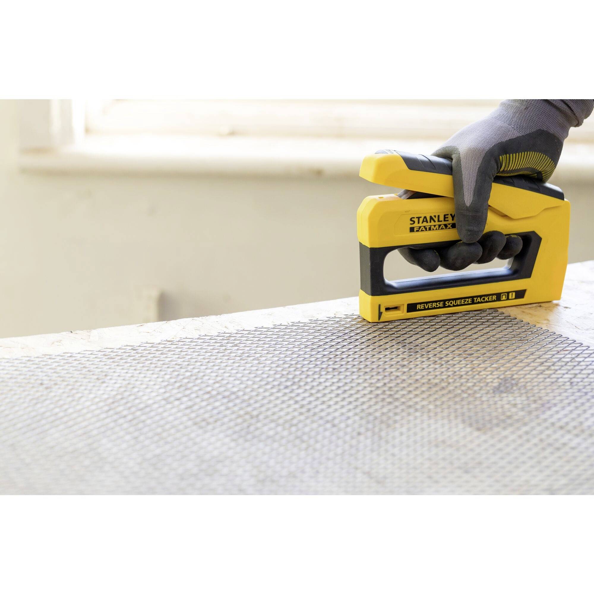 A hand wearing a glove is using a yellow staple gun to secure a wire mesh on a wooden table.