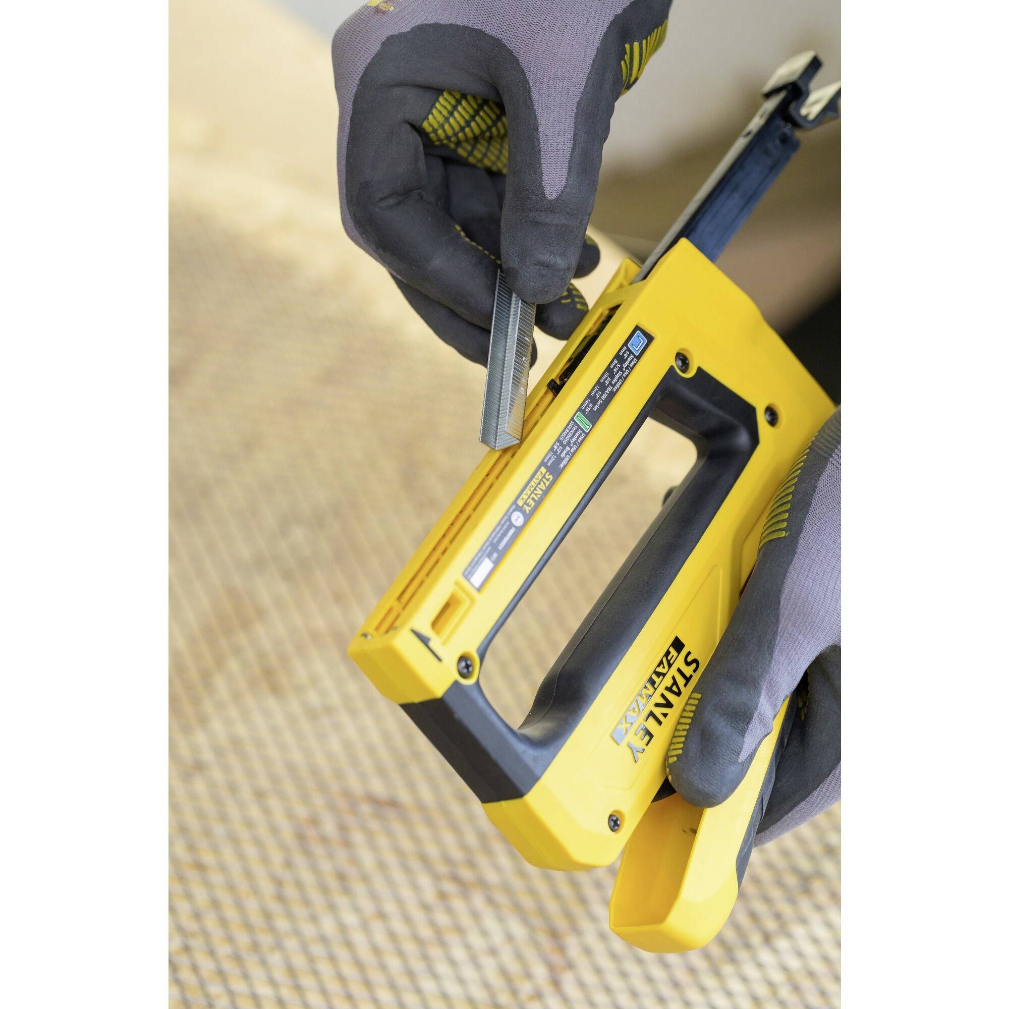 A person wearing work gloves is loading staples into a yellow staple gun, which is positioned on a carpet, prepared for DIY work.