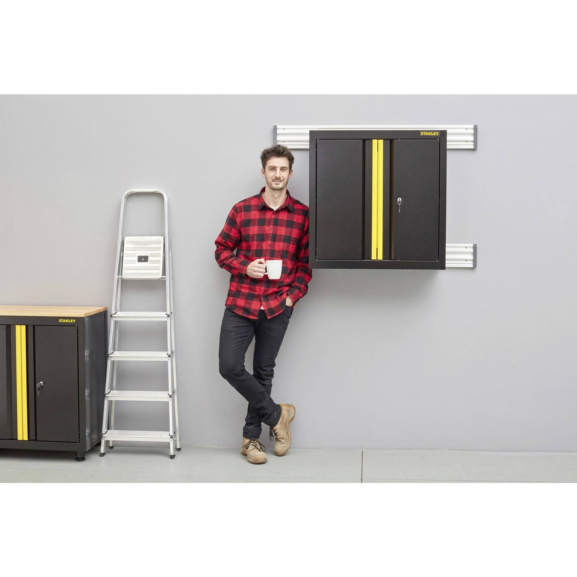 A man in a checked shirt is leaning against a ladder, holding a mug, and standing next to black cupboards with yellow stripes on the wall.