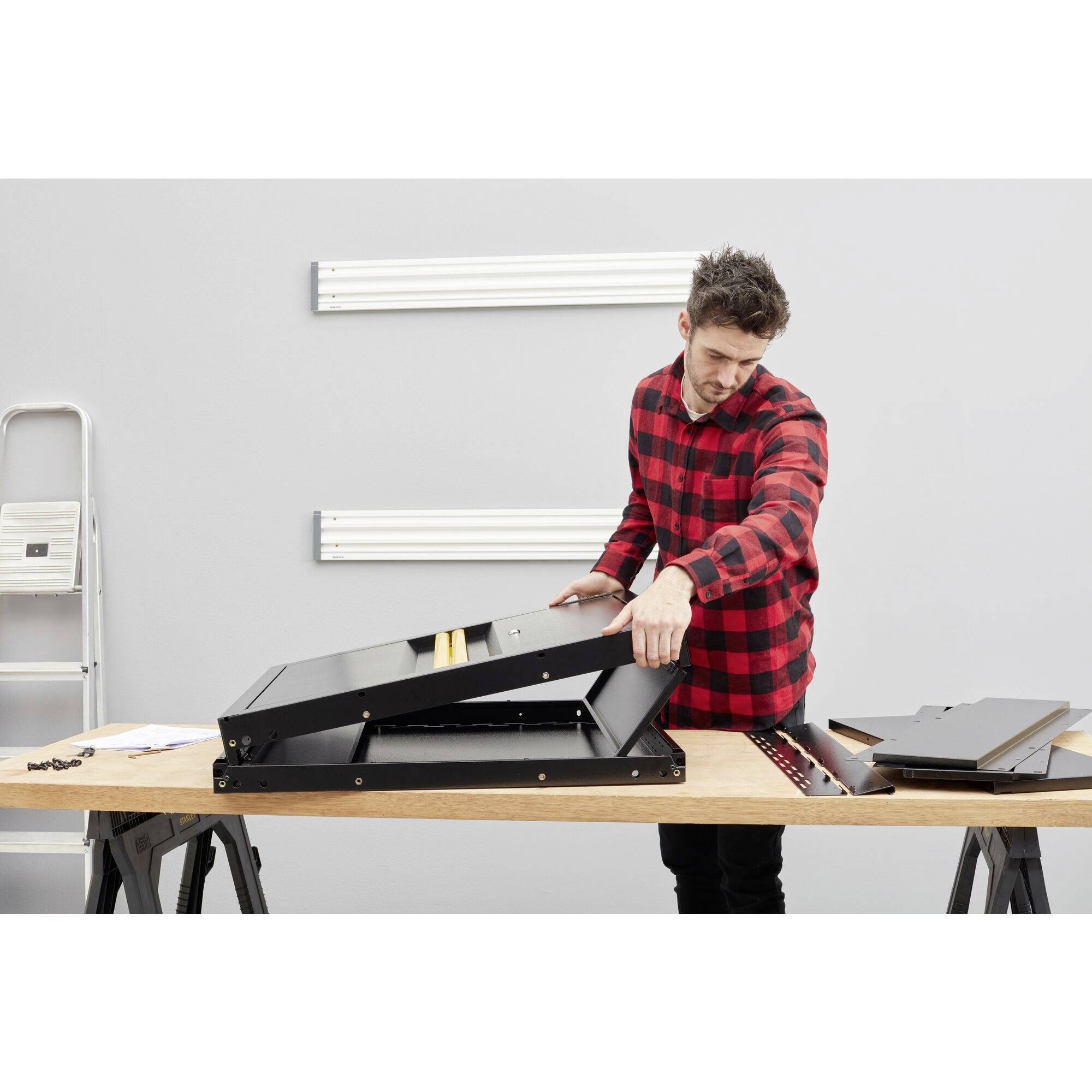 A man in a red shirt is assembling a black table on a workbench. Ladders and tools are visible in the background.