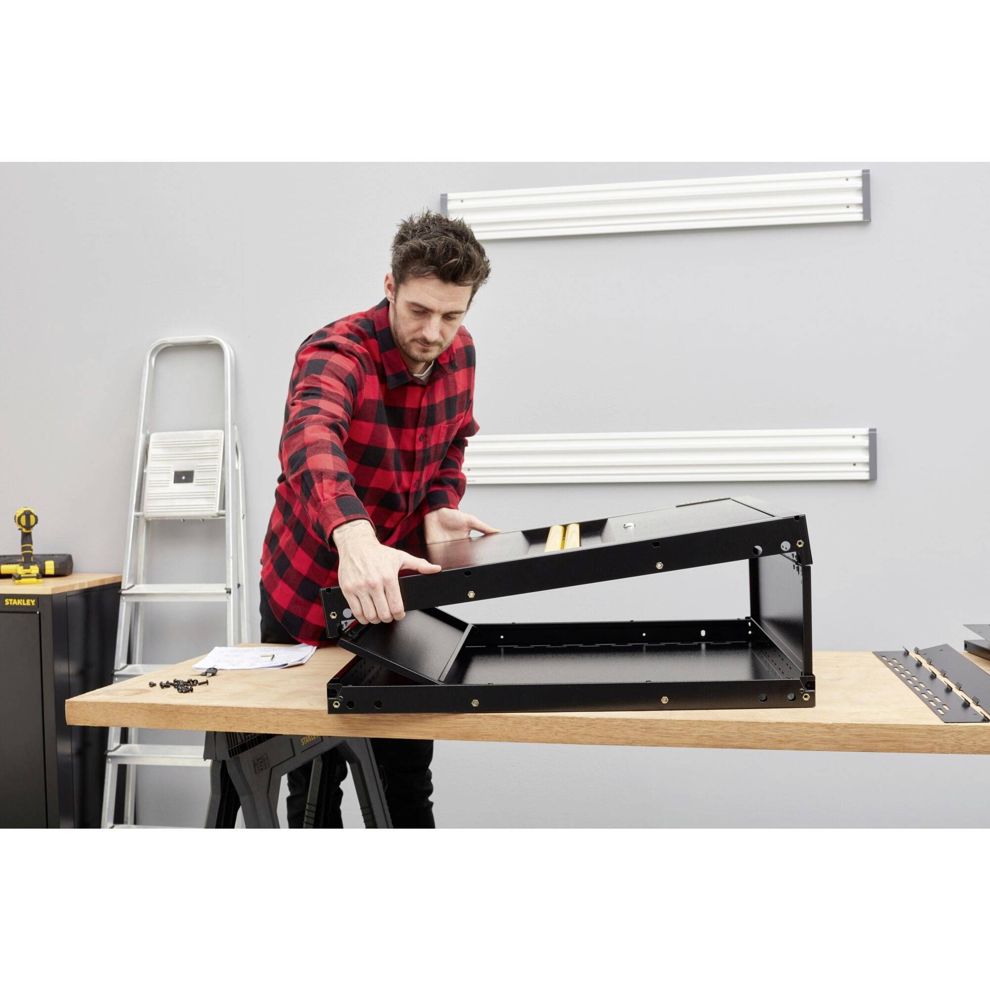 A person wearing a checked shirt is assembling a black metal frame on a table in a workshop environment with a ladder in the background.