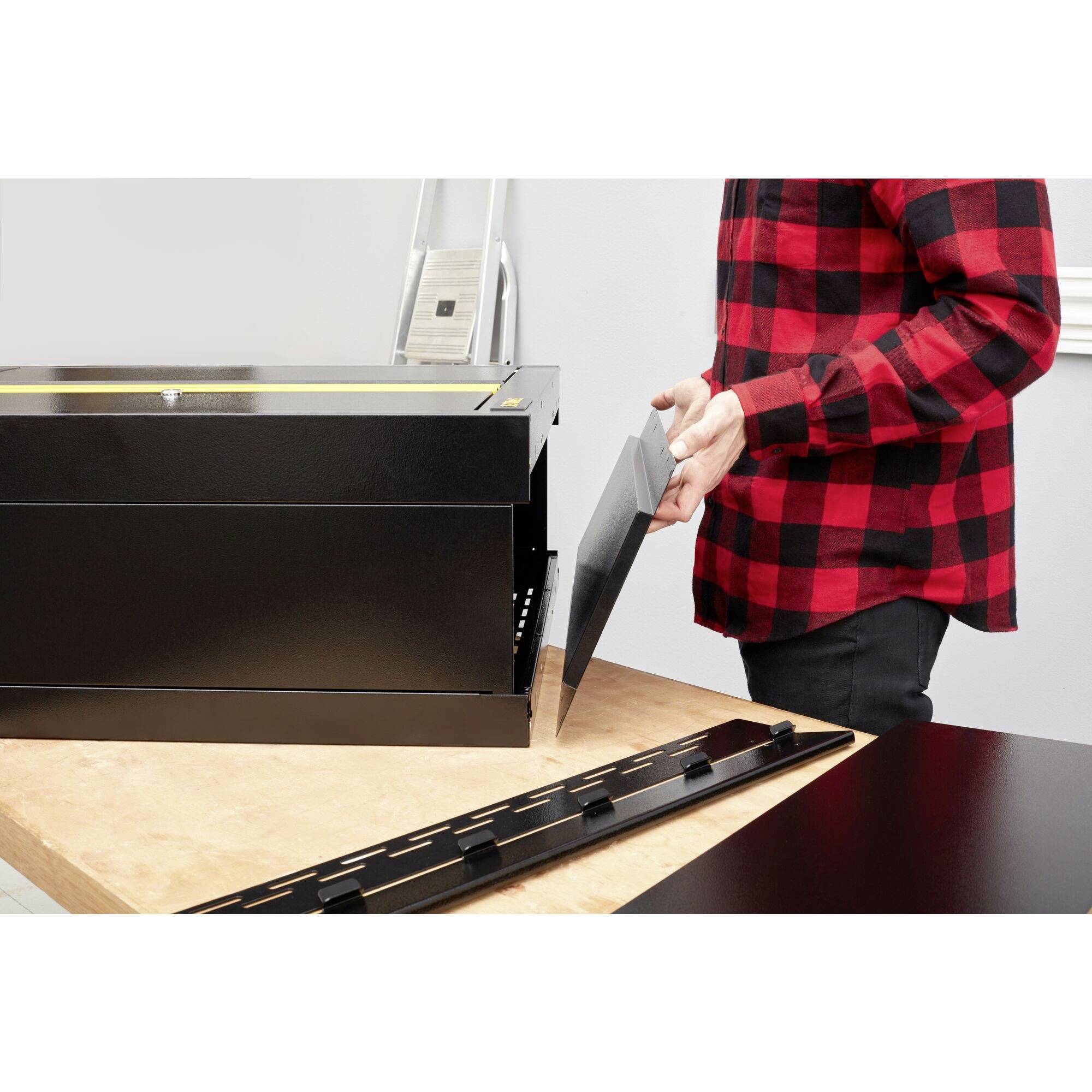 A person wearing a red and black checked shirt is assembling a black piece of furniture on a wooden table. A ladder is visible in the background.