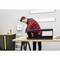 A person wearing a red and black checked shirt is assembling a black piece of furniture on a workbench in a workshop setting.