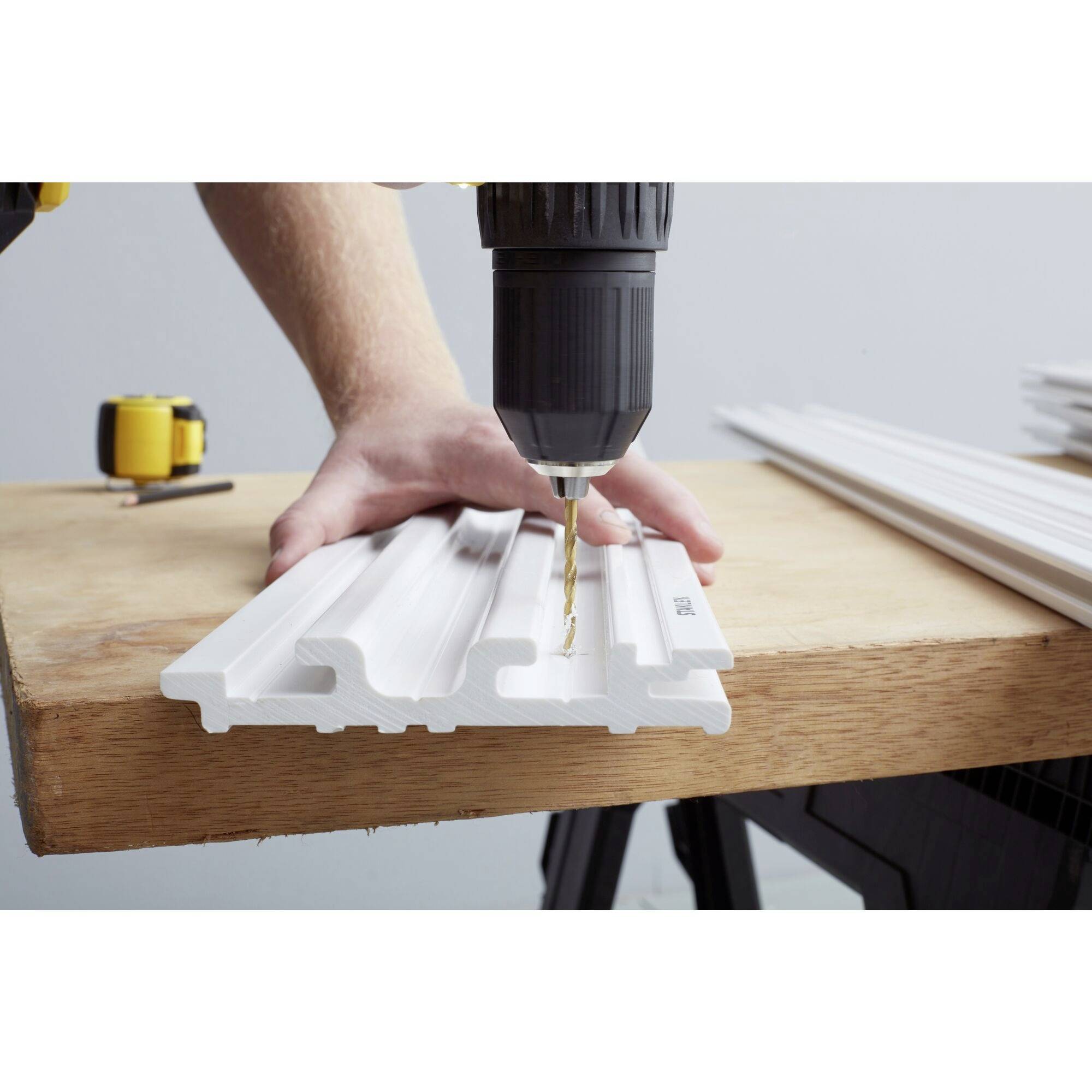 A person is drilling holes into a white plastic strip using a drill, which is lying on a wooden table. Additional materials are visible in the background.