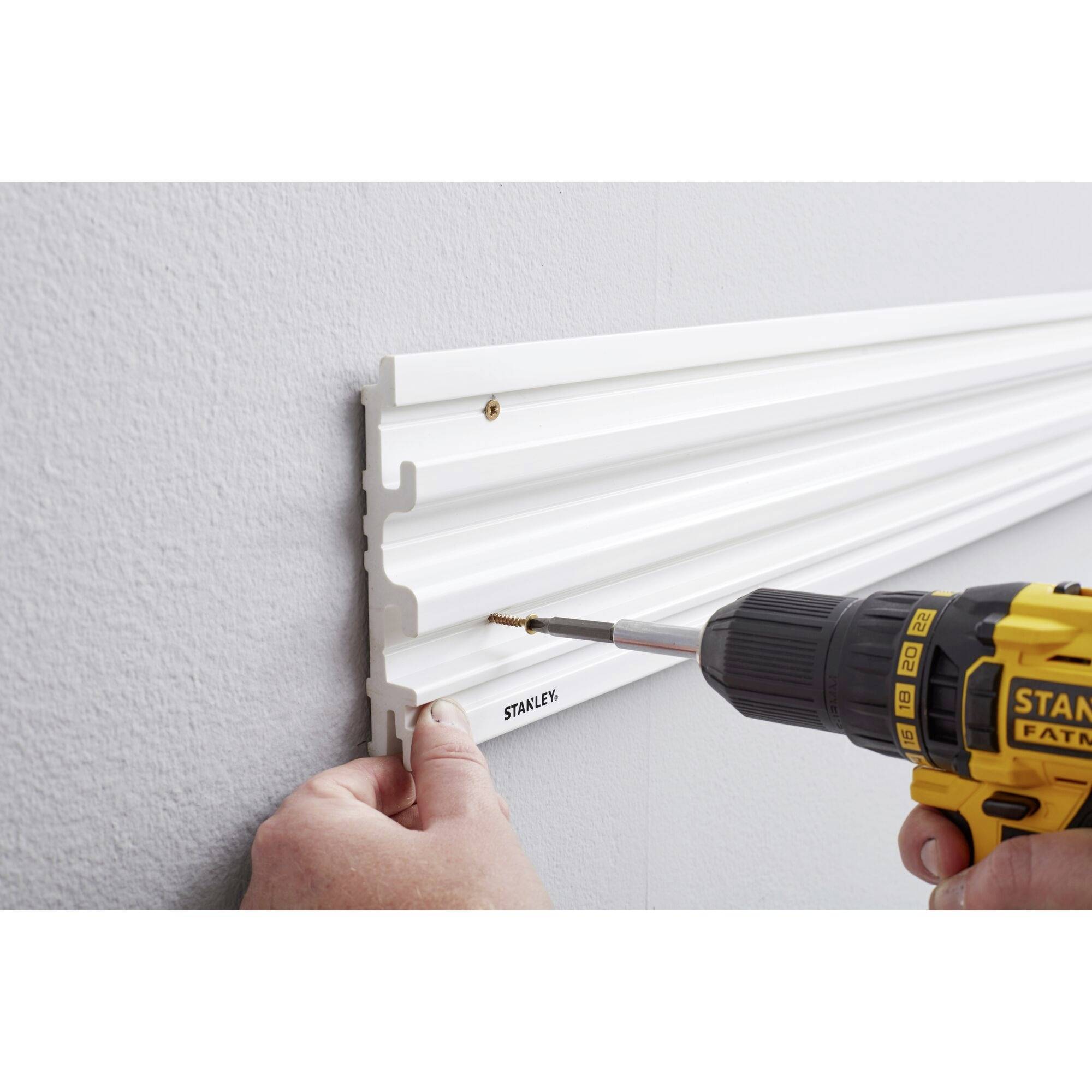 A person is securing a white plastic strip to a grey wall using a yellow cordless drill.