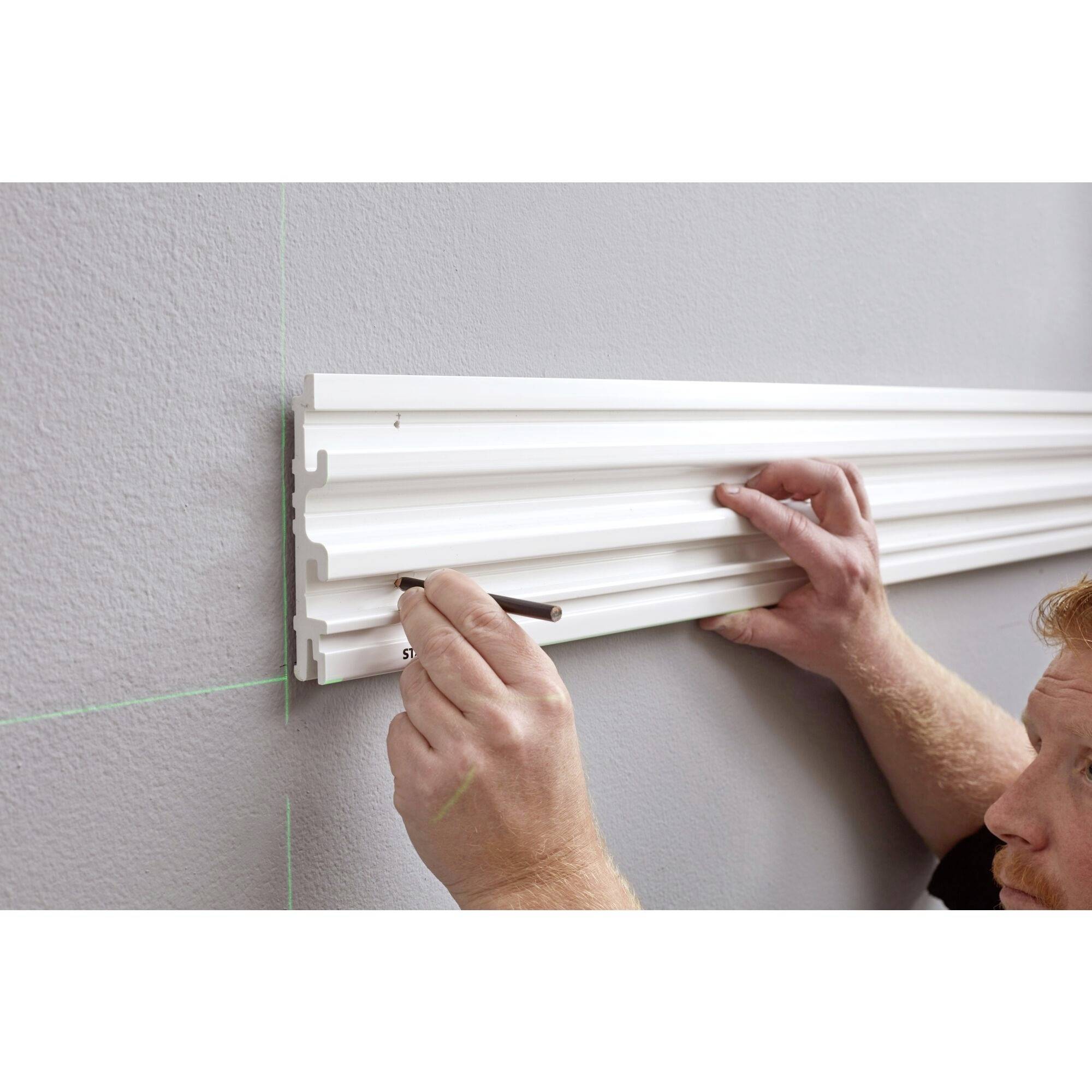 A person is installing a white wall cladding. They are holding the panel with their right hand and marking the wall with a pencil in their left.