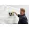 A man is hanging a black metal basket on a white, ribbed wall. The basket contains tools such as a tape measure.
