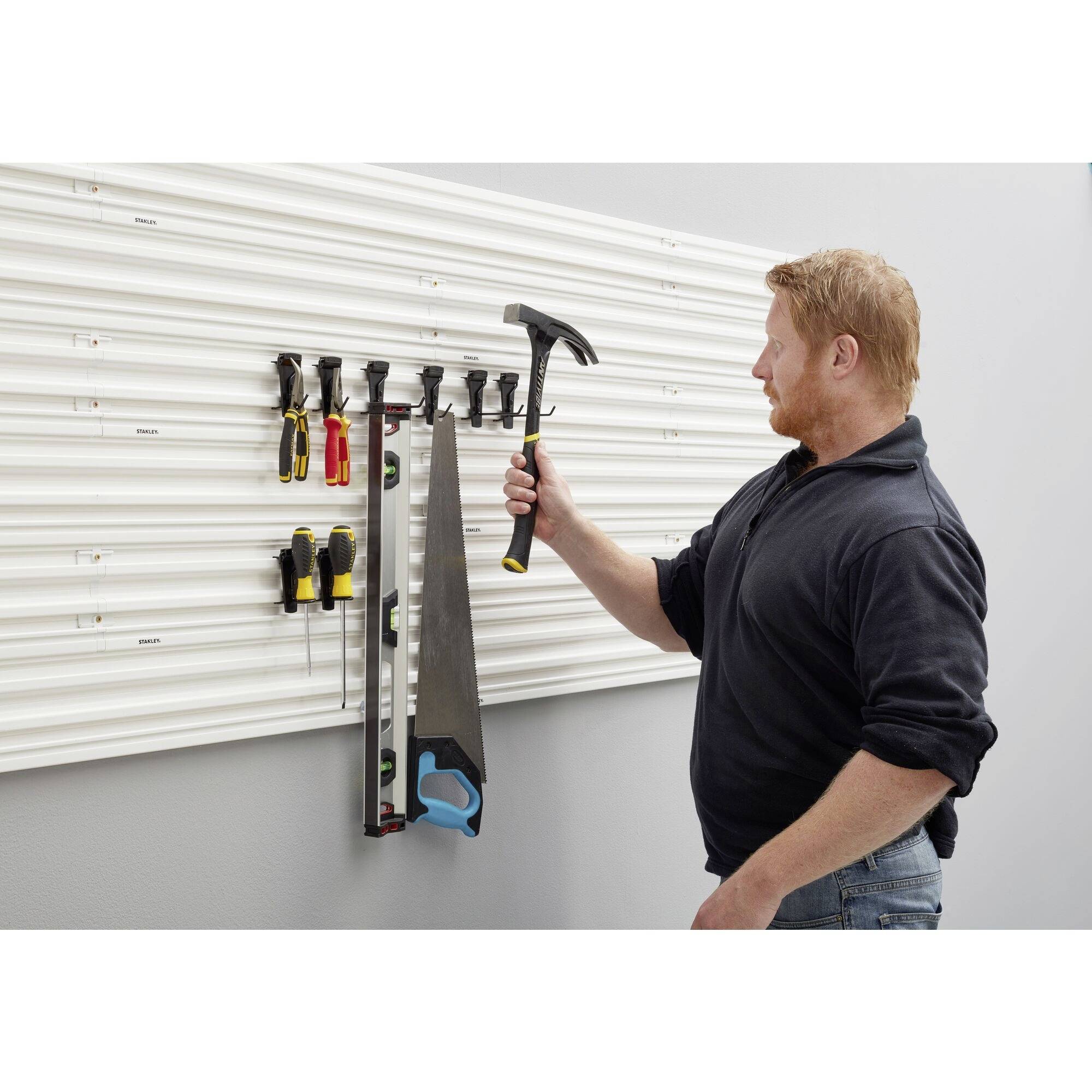 A man is hanging a hammer on a wall bracket that organises various tools, including screwdrivers and a saw.