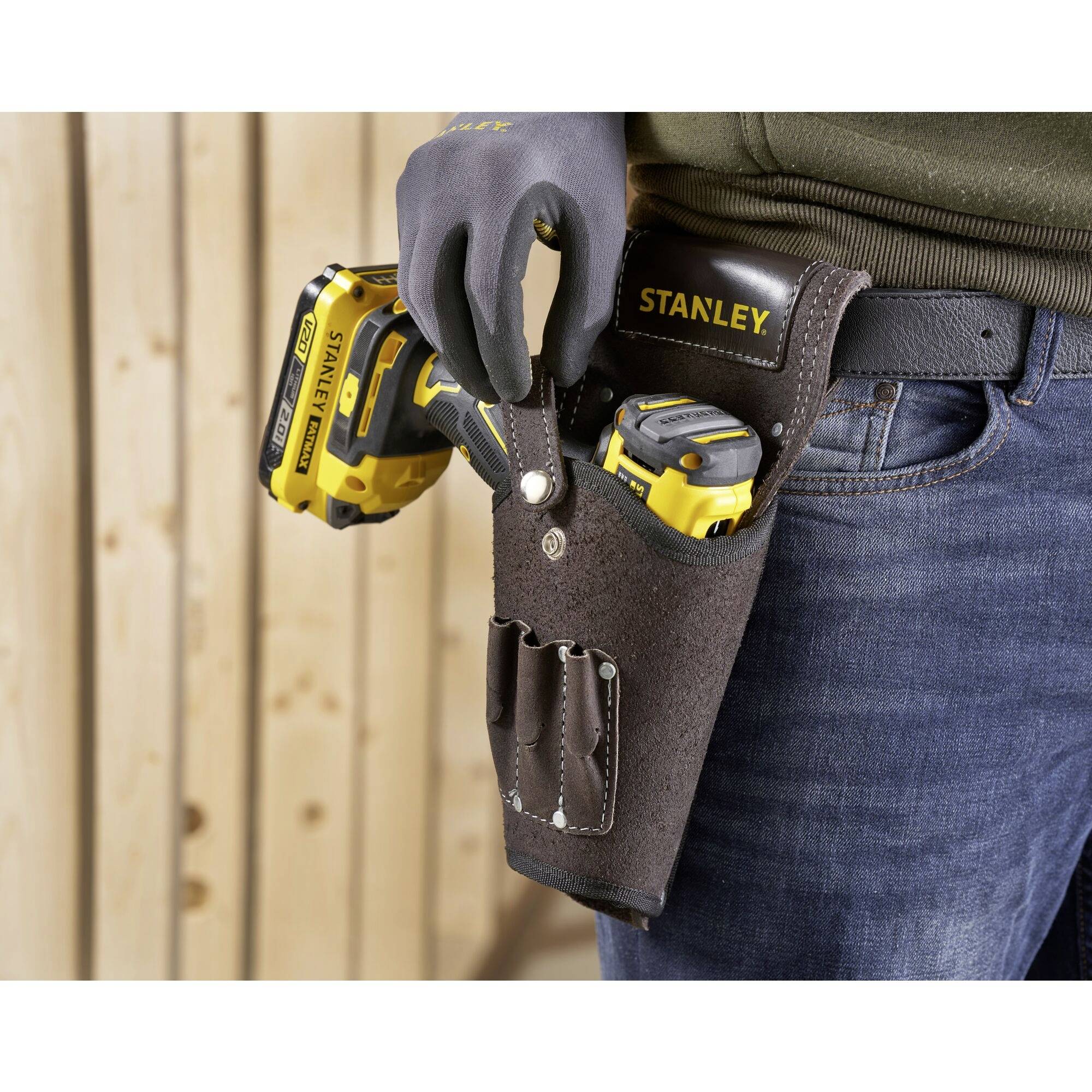 A person is standing in front of a wooden wall, wearing a black tool holster with a yellow cordless drill attached to their right hip.
