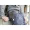 A person is wearing blue work trousers and a grey jumper, leaning against a wall. A tool belt is hanging from their trousers.