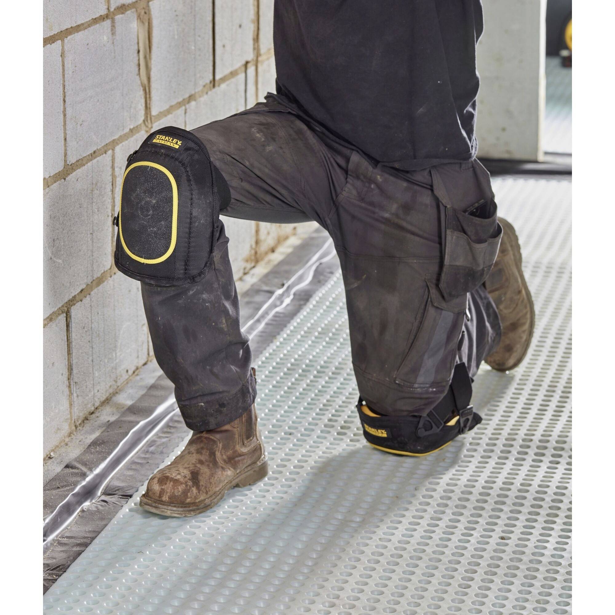 A person is kneeling on a building site. They are wearing knee pads and workwear. The ground is covered with a plastic sheet.