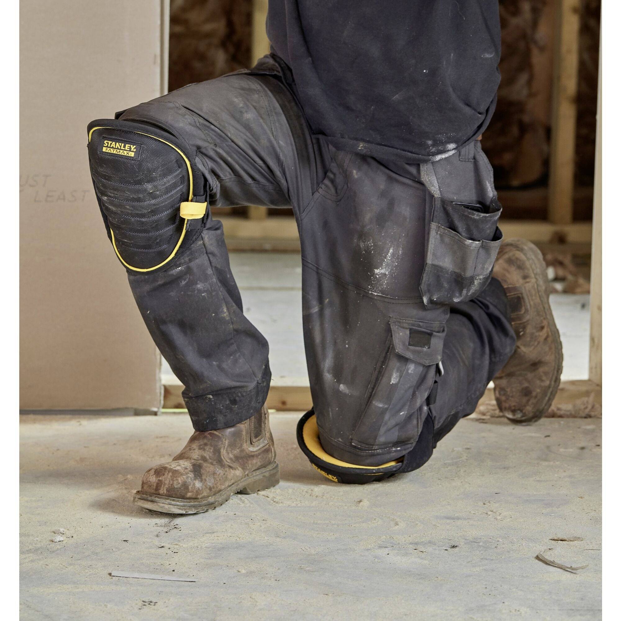 A person is kneeling on the ground at a construction site, wearing robust work boots, black trousers with tool pockets, and a knee pad.
