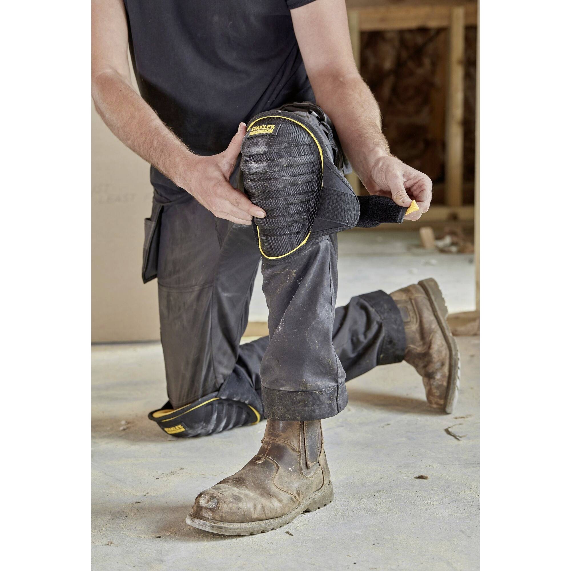 A person is kneeling on a building site, wearing knee pads. The ground is dusty and the person is wearing work clothing and boots.
