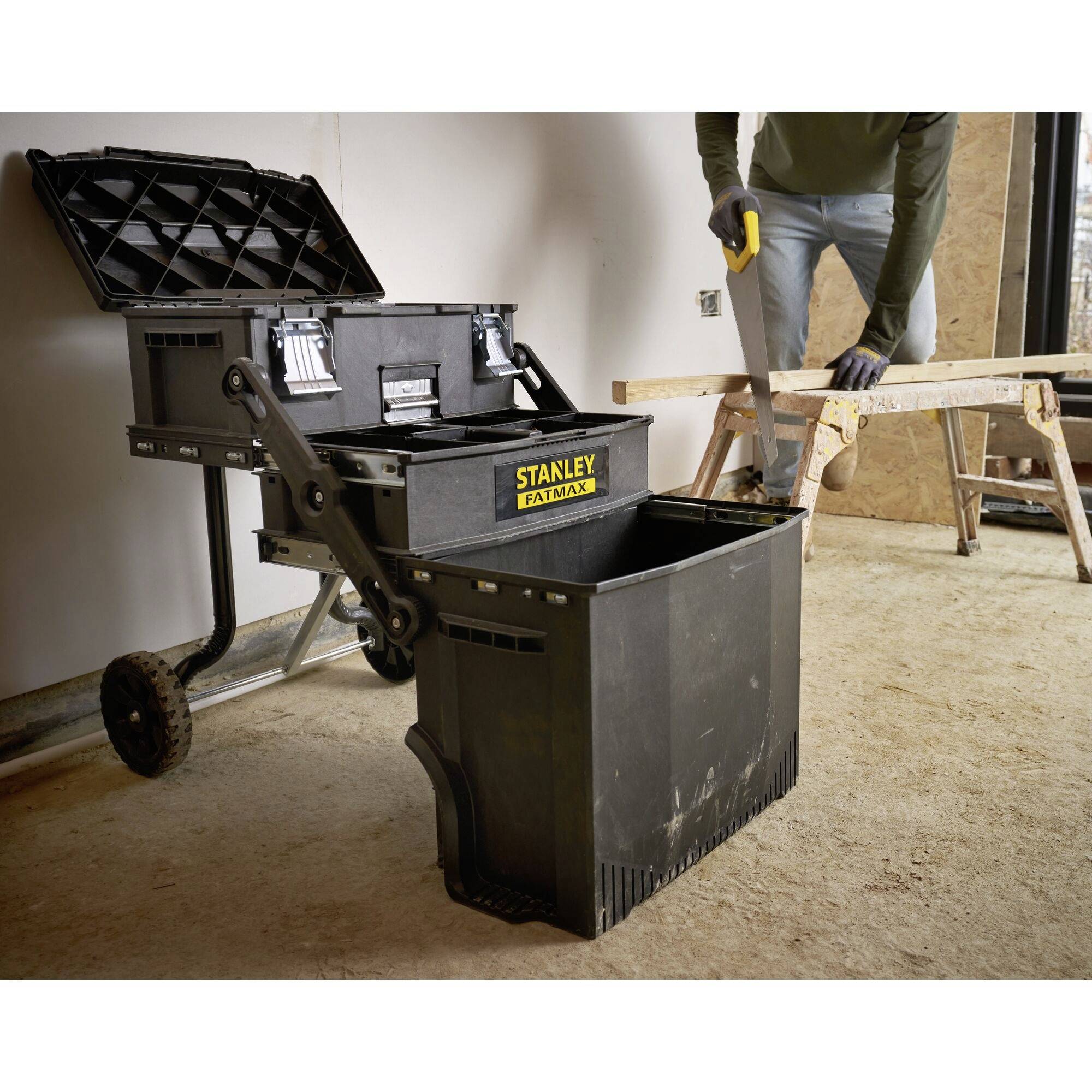 An open toolbox on wheels stands in a workshop. A person in the background is working with a piece of wood on a workbench.