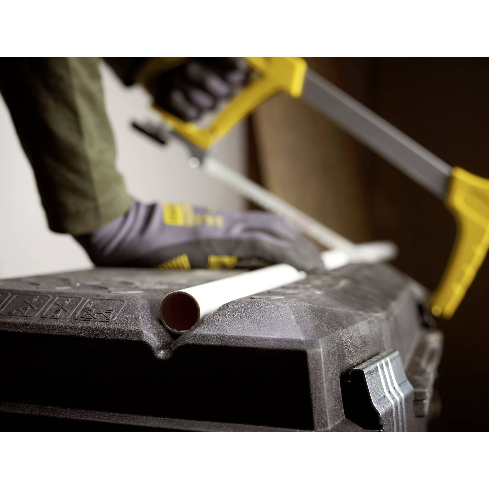 A person is sawing a white pipe with a hacksaw while wearing grey gloves. The environment is a workshop.