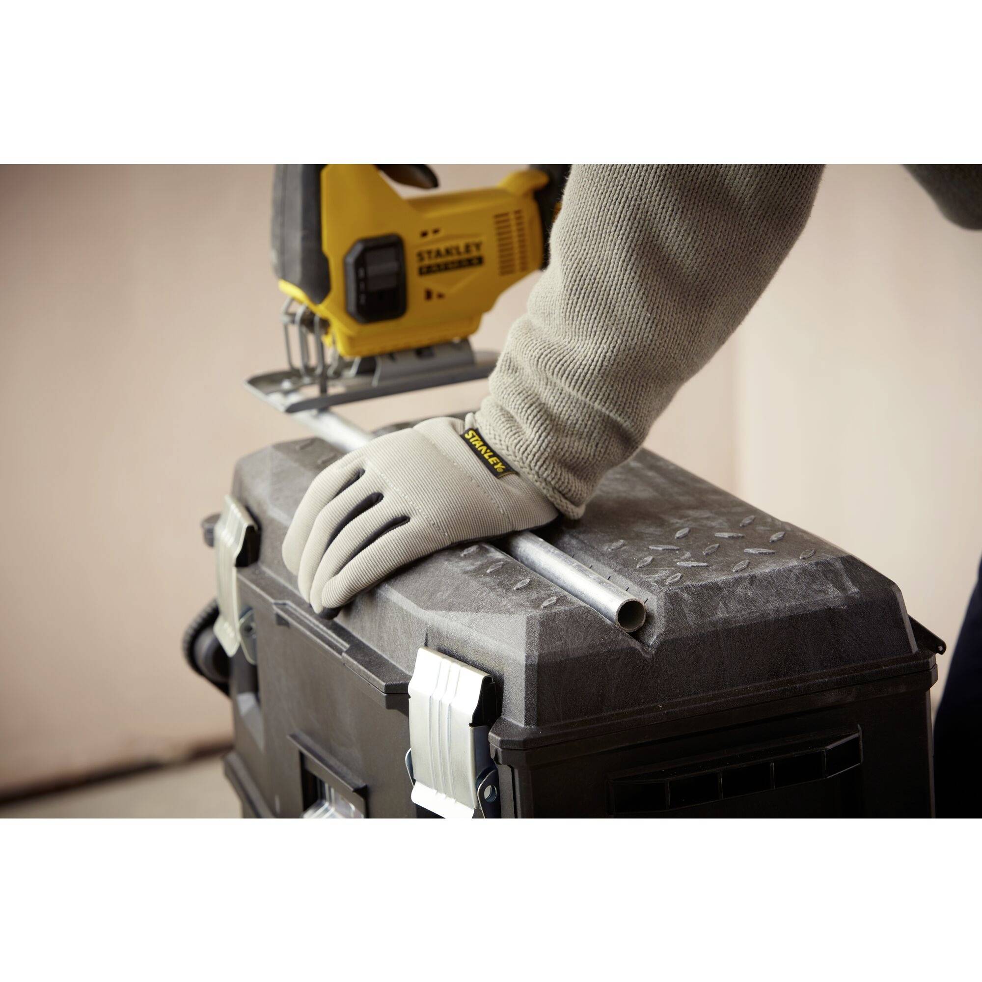 A person wearing a grey glove is sawing a metal pipe with a yellow jigsaw, which is resting on a black crate.