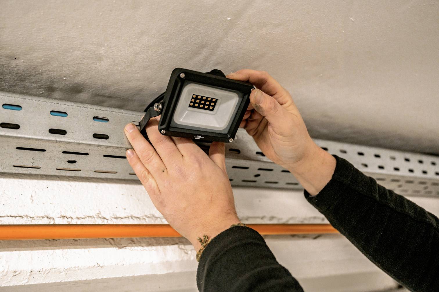 A person is installing an LED floodlight on a metal bracket on the ceiling.