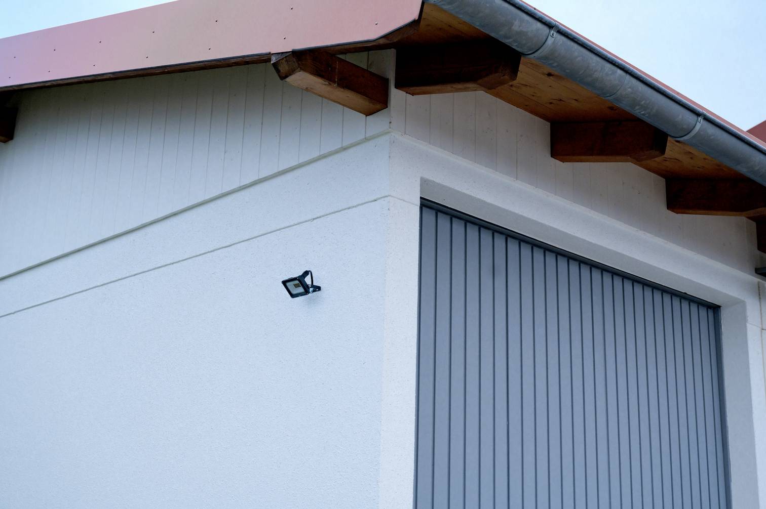 A modern house roof with red tiles and a guttering, next to a grey garage door and a security camera on the white wall.