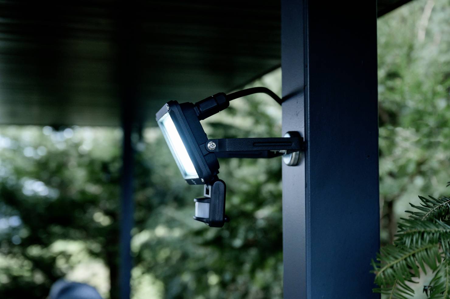 A LED floodlight is mounted on a post outdoors and is illuminating to the side to light up a dark environment. Trees can be seen in the background.