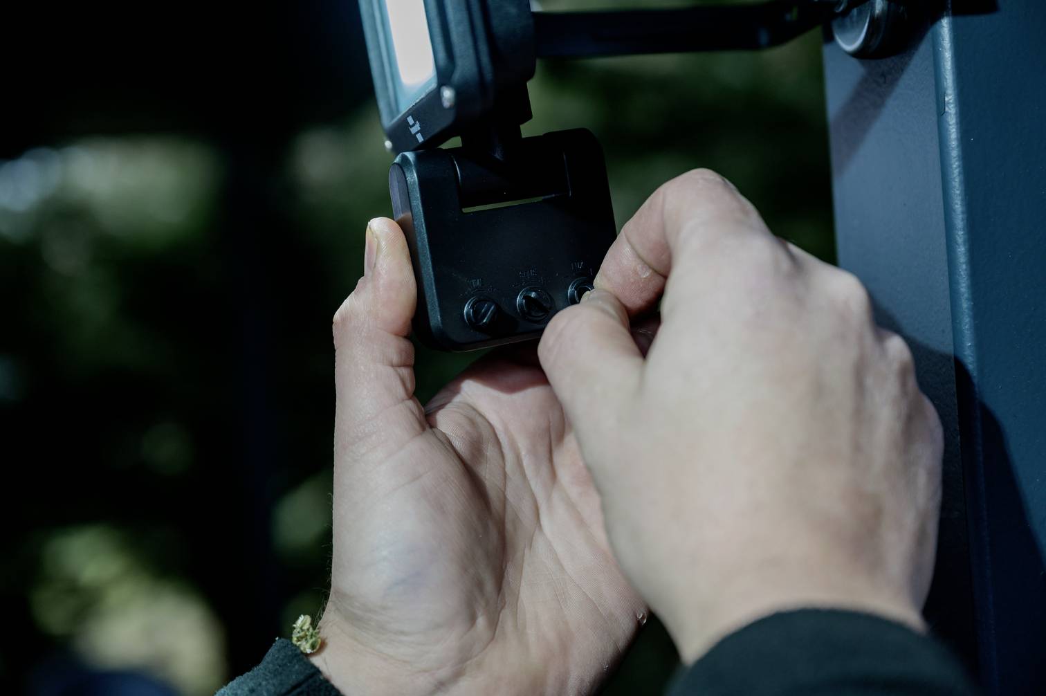 Two hands are adjusting a small black device on a grey object. The surroundings are blurred and dimly lit.