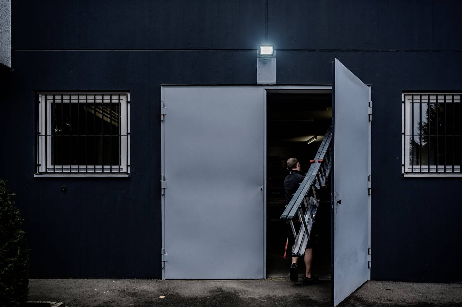 A person is carrying a ladder through a partially open metal door into a dark grey building. Barred windows flank the door.