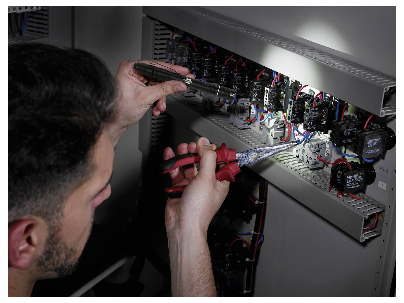 Technician wiring and inspecting an electrical control panel with a screwdriver and pliers, surrounded by relays, switches, and cables.<br>