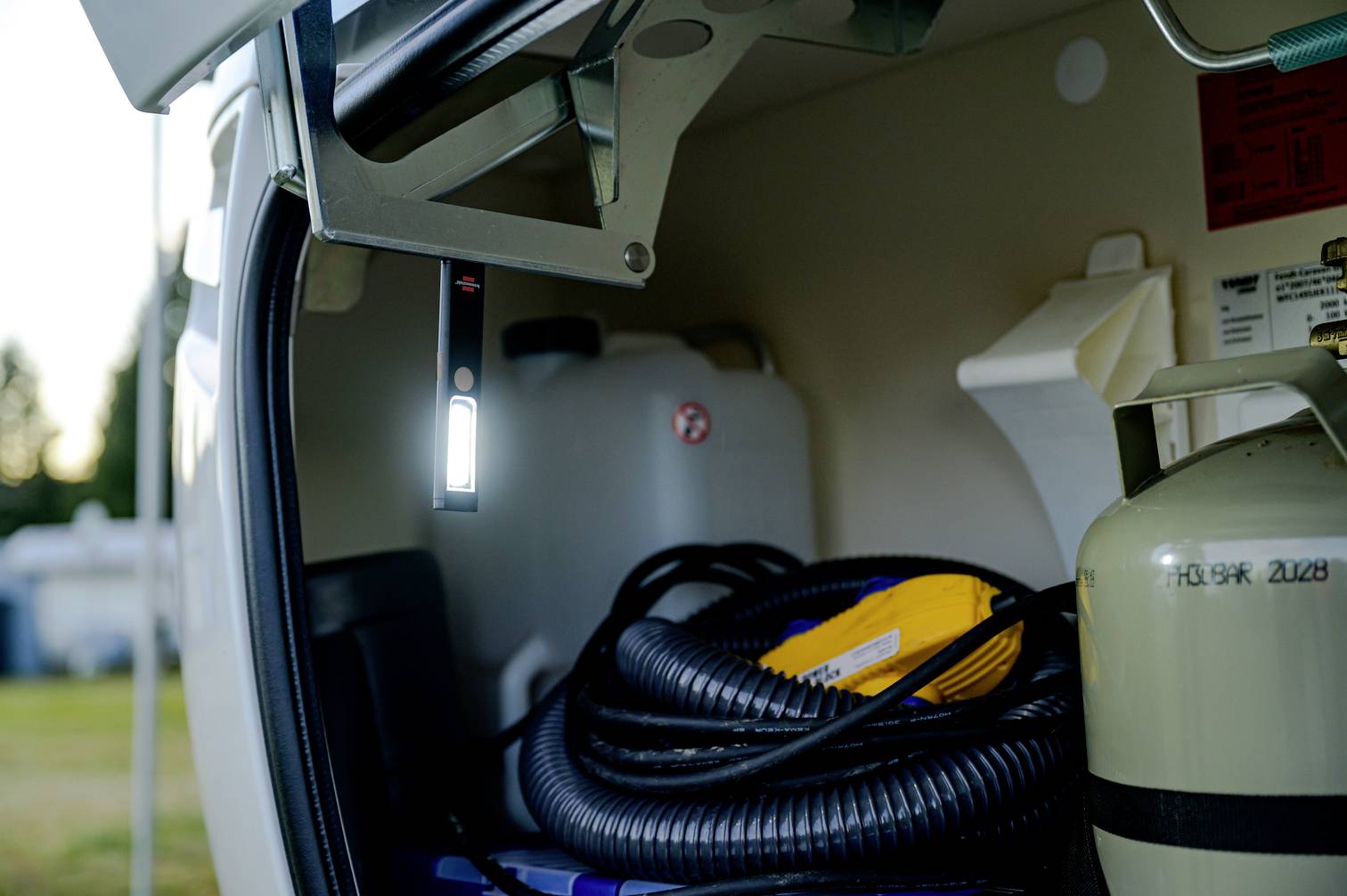 'A vehicle storage compartment with a gas cylinder, hoses and water container. A light illuminates the interior.'