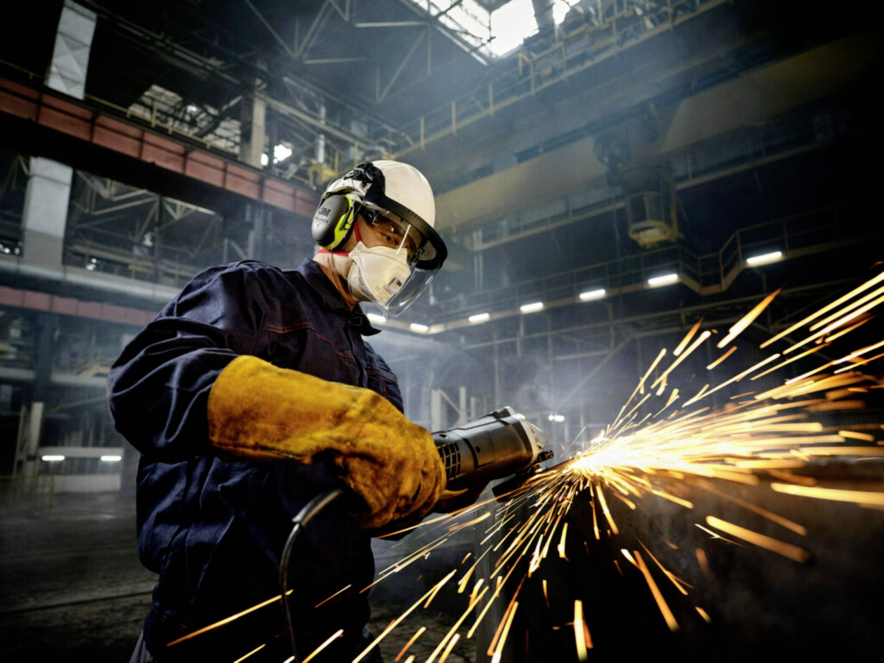 A worker in protective clothing grinds metal in a factory hall. Sparks fly from the grinding point.