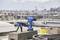 A blue pliers set lies on a roof next to concrete slabs and pipes. A blurred urban landscape is visible in the background.