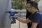 A man is installing a part on a wall using a blue power tool. He is wearing safety glasses and work gloves.