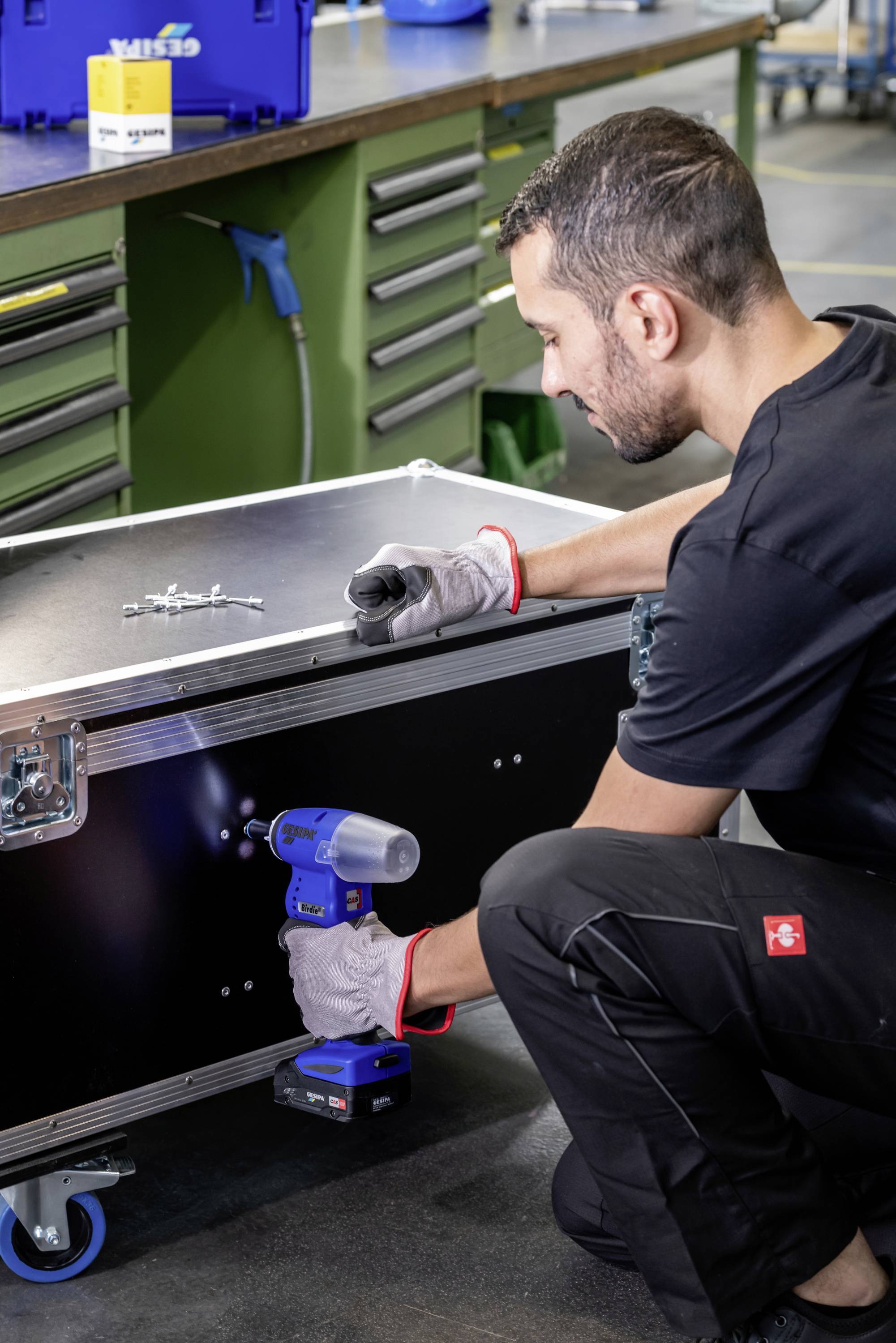 A man in workwear is using an electric screwdriver to repair a black wheeled box in a workshop.