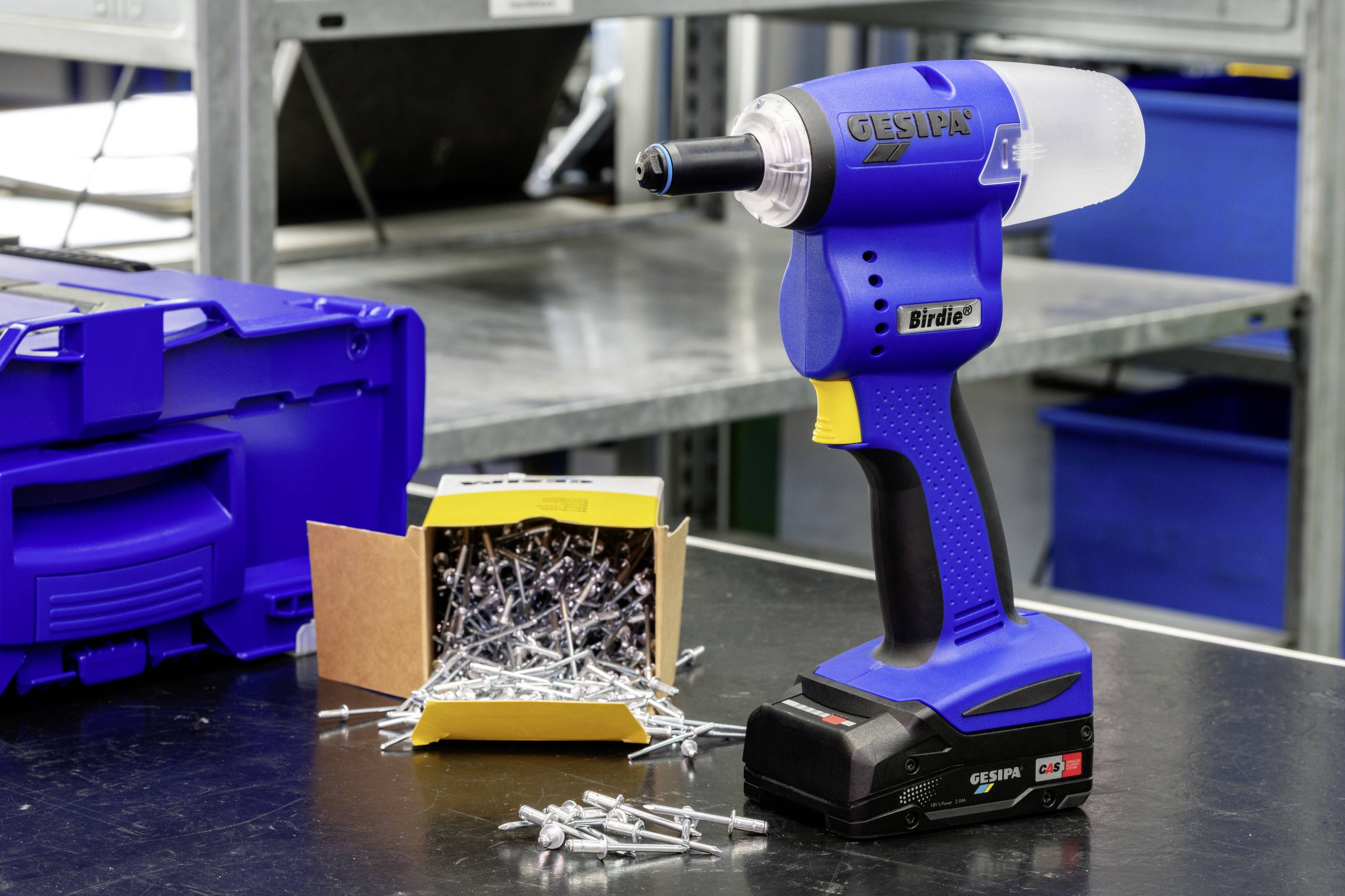 A blue and yellow cordless drill sits on a table next to an open box of rivets. Metal shelving is visible in the background.