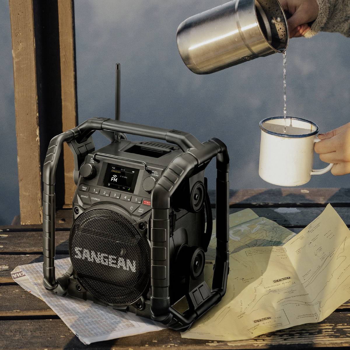 A hand pours coffee into an enamel mug next to a radio. Cards are laid out on the wooden table, suggesting an outdoor scene.