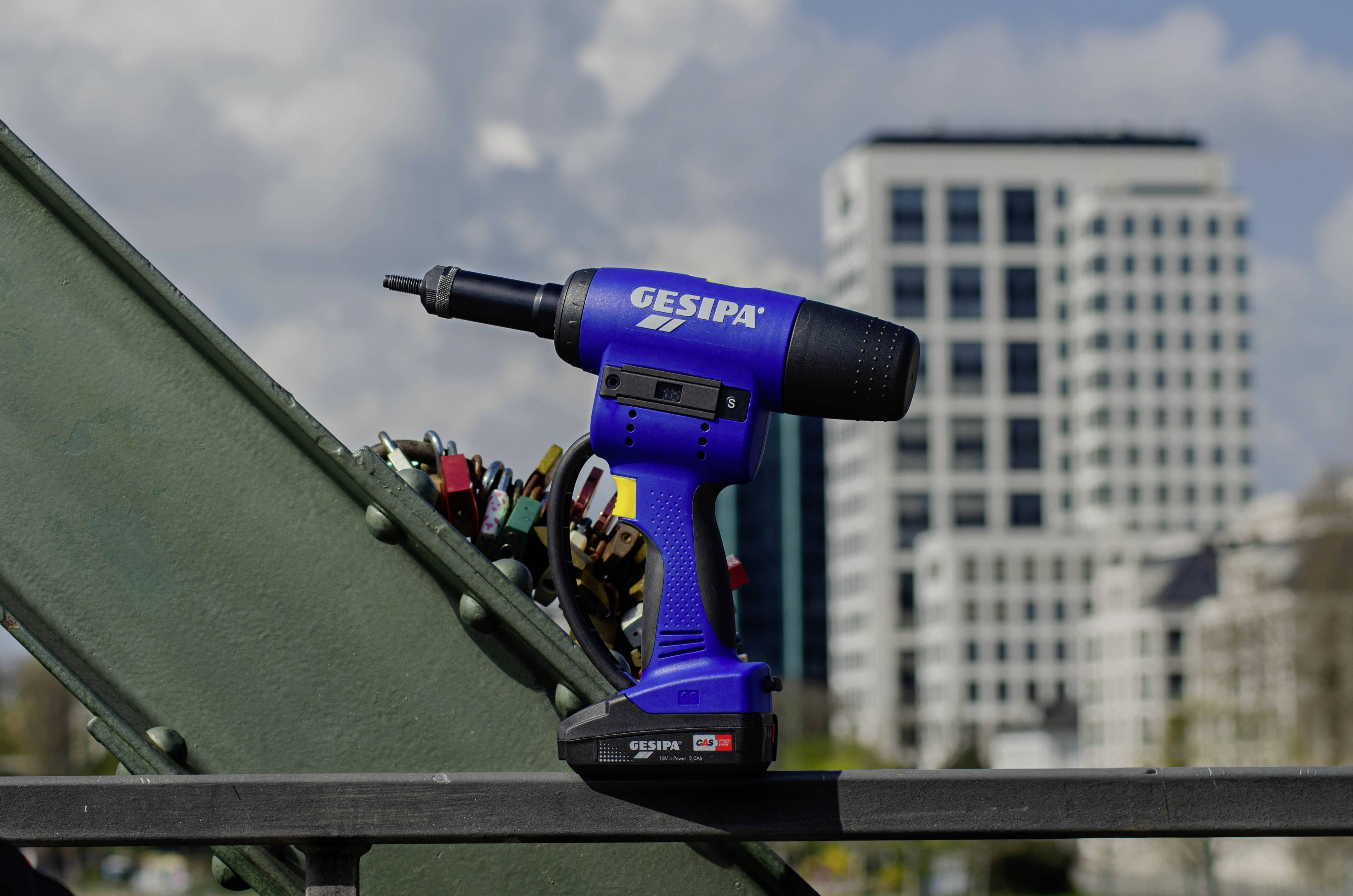A blue Gesipa cordless drill sits on a steel beam. A modern office building is visible in the background.