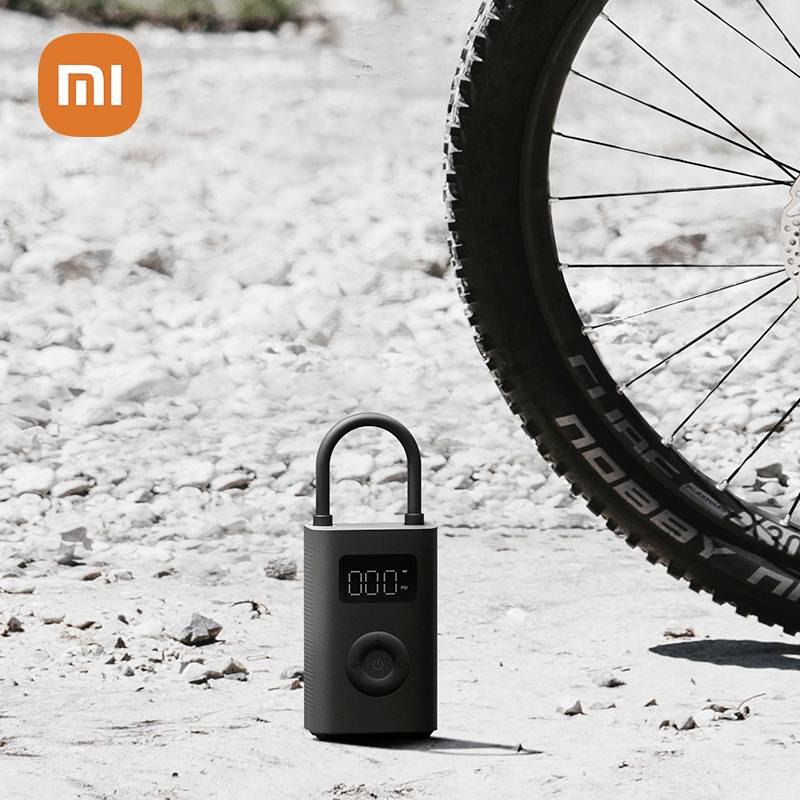 A portable black air pump stands next to a bicycle tyre on a gravelly surface. An orange Mi logo is positioned in the top left corner.